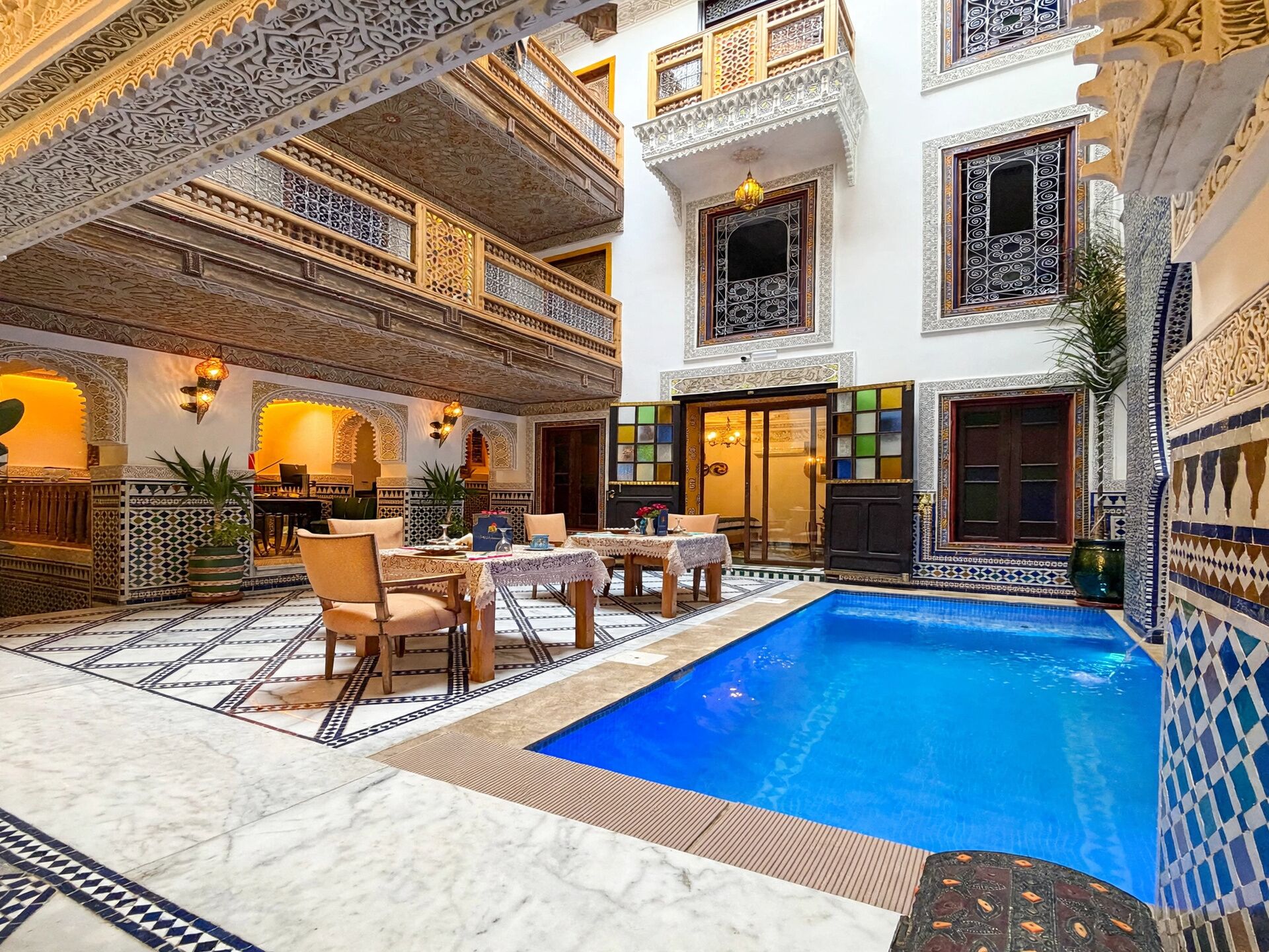A luxurious indoor pool area with intricate tile work, a dining setup, and a multi-level balcony with ornate railings.