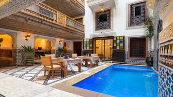 A luxurious indoor pool area with intricate tile work, a dining setup, and a multi-level balcony with ornate railings.