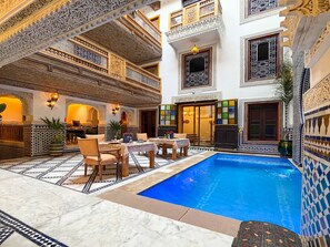 A luxurious indoor pool area with intricate tile work, a dining setup, and a multi-level balcony with ornate railings.