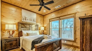 Ein Schlafzimmer einer Holzhütte mit einem Bett, Nachttischen, einem Deckenventilator und einem großen Fenster.