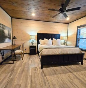 Un dormitorio con piso de madera, una cama con cabecera oscura, dos mesitas de noche con lámparas, un escritorio y un ventilador de techo.