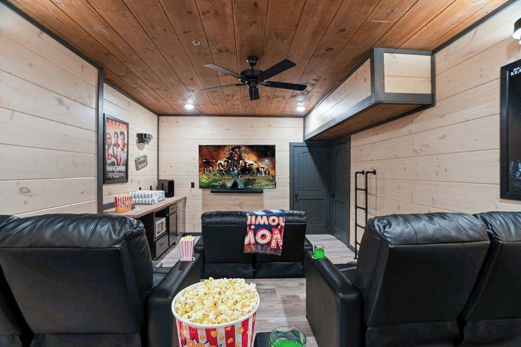 Una sala de cine en casa con paredes de madera, ventilador de techo, cartel grande, sofás de cuero negro, palomitas de maíz y una consola de videojuegos.