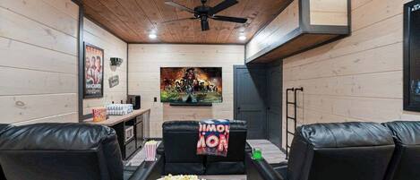 Una sala de cine en casa con paredes de madera, ventilador de techo, cartel grande, sofás de cuero negro, palomitas de maíz y una consola de videojuegos.