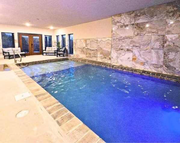 Una piscina interior con paredes de piedra, suelo de baldosas y zona de estar con sillas y una mesa.