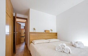Une chambre d’hôtel avec une tête de lit en bois, un lit recouvert de draps blancs, une armoire en bois et une petite étagère avec deux lampes.