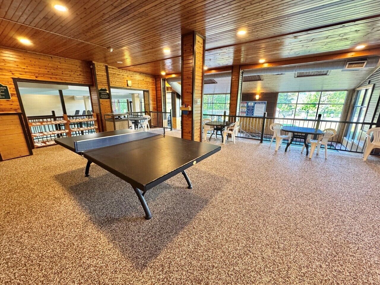 Une table de ping-pong se trouve dans une pièce aux murs en bois et au sol recouvert de moquette, entourée de chaises.