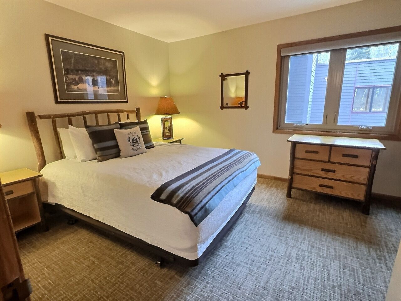 A bedroom with a bed, bedside table, lamp, dresser, and a window.