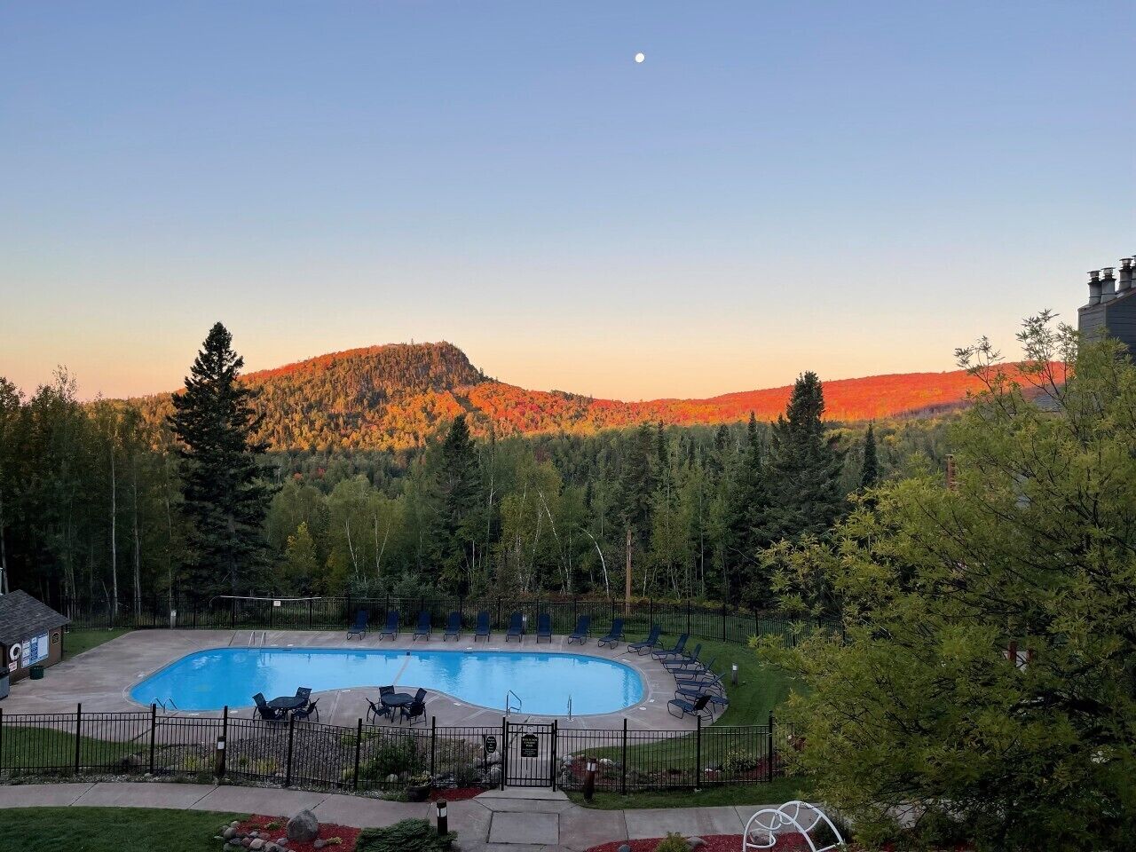 A swimming pool with lounge chairs, surrounded by a fence, with a forested hill in the background.