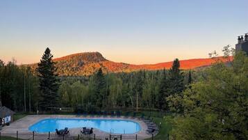 A swimming pool with lounge chairs, surrounded by a fence, with a forested hill in the background.