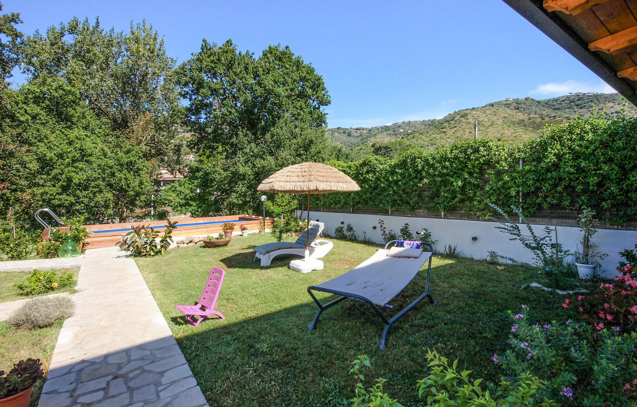 Um quintal com piscina, cadeiras de descanso, um guarda-sol de palha e um caminho de pedra.