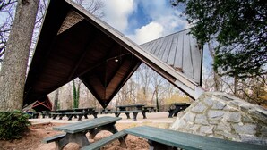 Een overdekte picknickplaats met banken en picknicktafels, omgeven door bomen en een stenen muur.