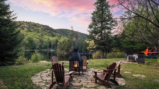Un foyer en pierre, entouré de fauteuils Adirondack, d’une table et d’un barbecue, le tout installé devant un paysage composé d’un lac et de collines boisées.