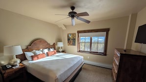 Um quarto com cama, mesas de cabeceira, abajur, cômoda, ventilador de teto, janela com vista e um quadro na parede.