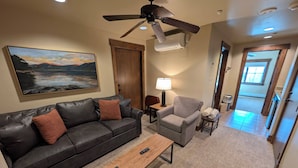 Uma sala de estar com sofá de couro, poltrona cinza, mesa de centro de madeira, quadro, ventilador de teto e luminária.