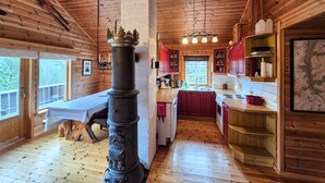 Een houten blokhut met een centraal gelegen open haard, een eethoek, een keuken met rode kastjes en een groot raam.