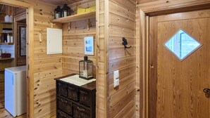 Een houten blokhut interieur met een deur, een kleine koelkast, een plank met spullen en een wandlamp.