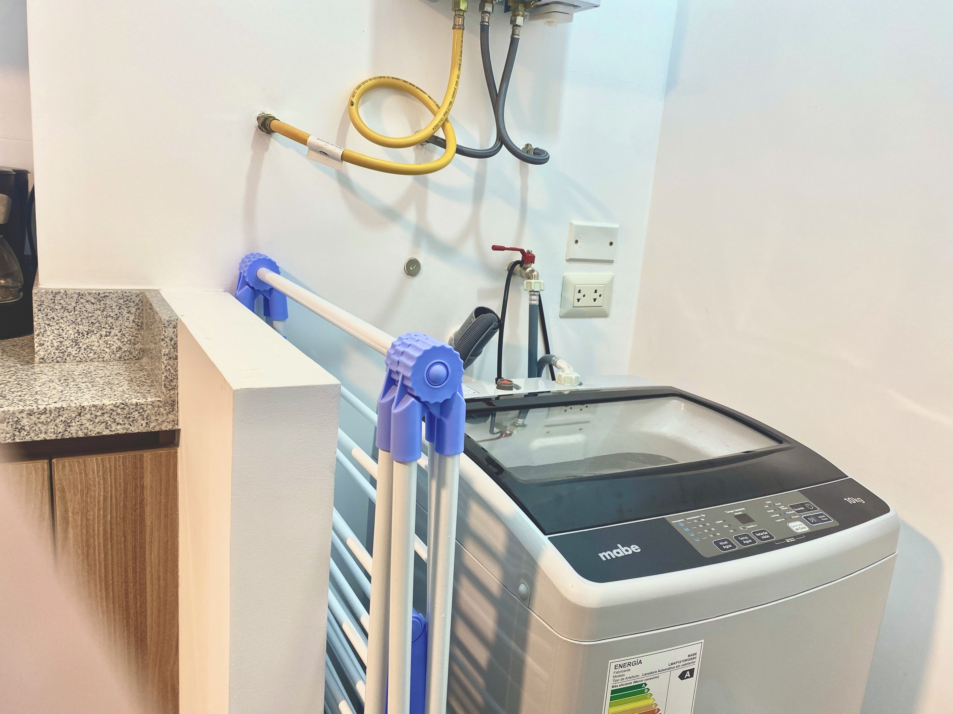 A modern, compact washing machine with a digital control panel, a water hose, and a power outlet on the wall.
