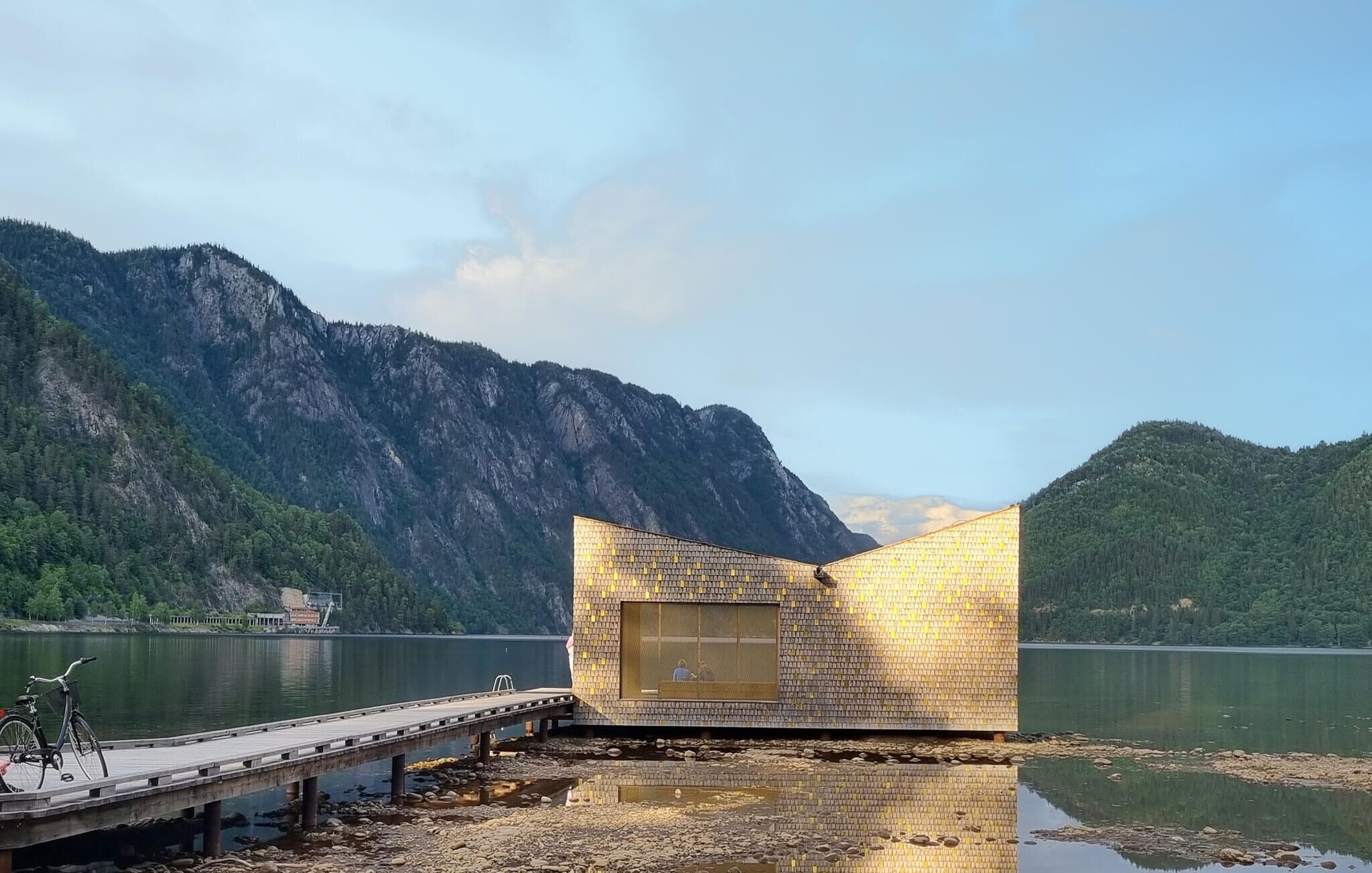 A modern building with a unique geometric design, situated by a calm lake with mountains in the background.