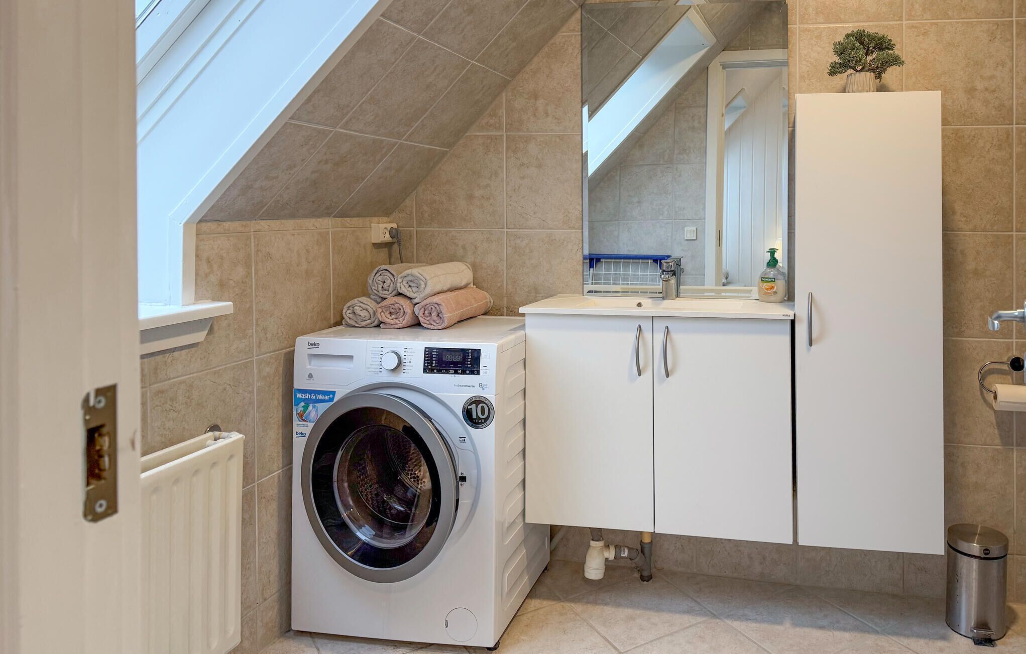 A compact laundry area with a front-loading washing machine, a drying rack with folded towels, a sink with a mirror above, and a cabinet with a trash bin.