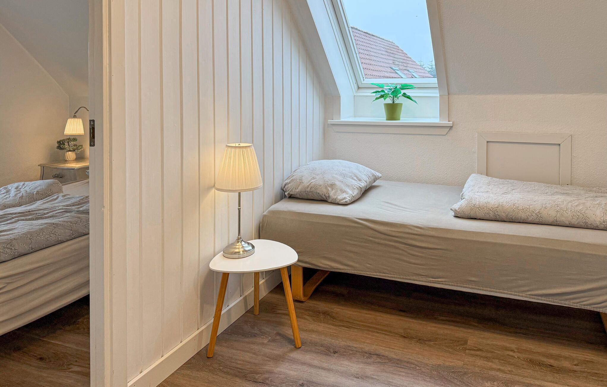 A small, cozy bedroom with a single bed, a bedside table with a lamp, a window with a plant, and wooden flooring.