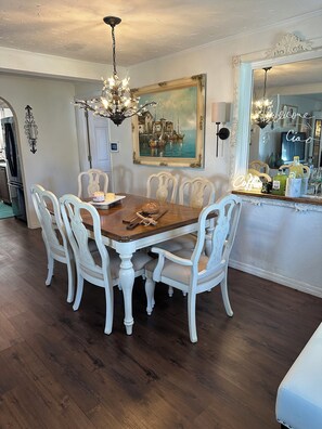 A dining area with a wooden table and white upholstered chairs, a chandelier, and a painting on the wall. - Welcoming spacious Beach Cottage in  Sarasota near Siesta Key  w/ Luxury Decor (Sarasota)