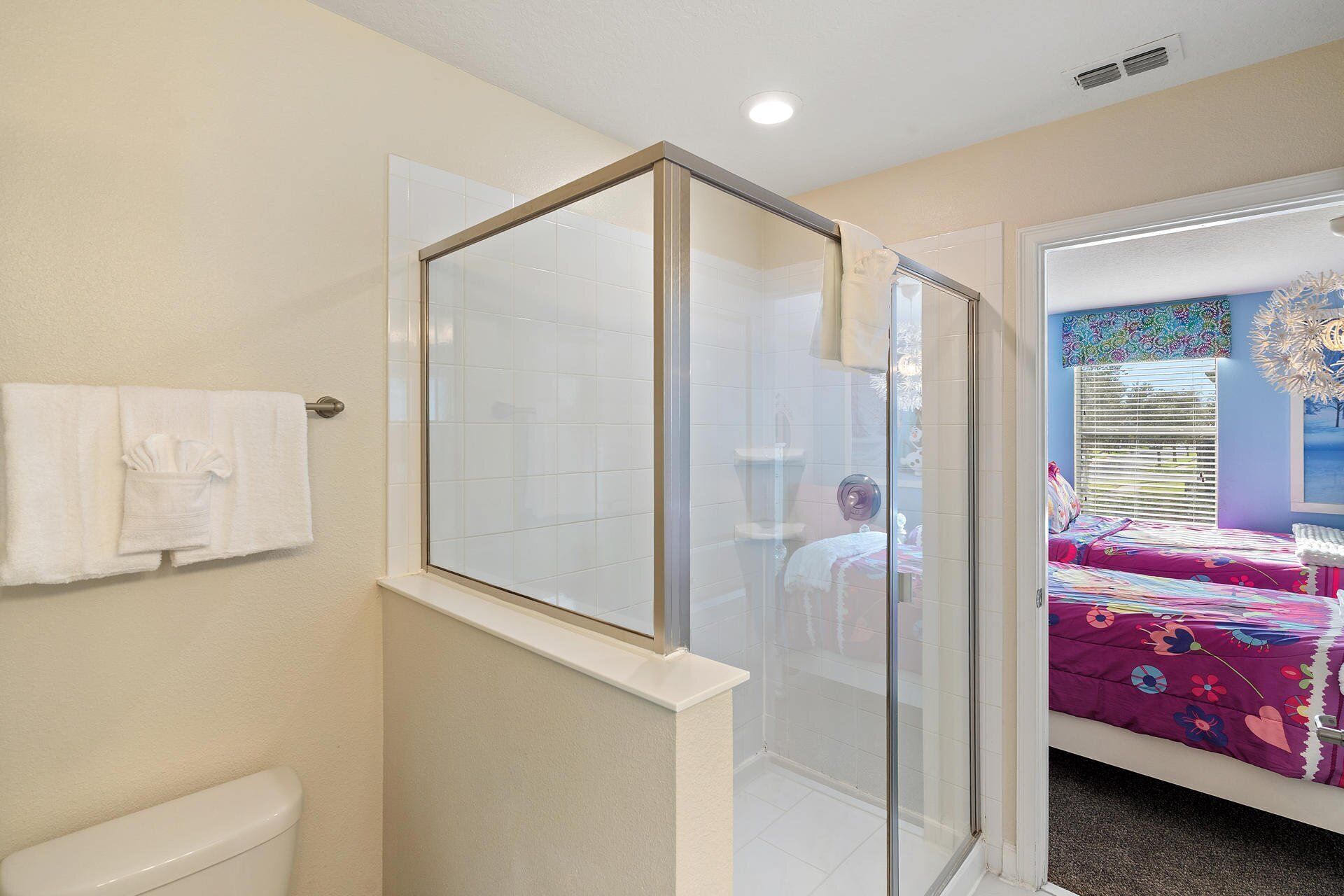 Un baño con ducha de vidrio, inodoro y cama con colcha colorida.
