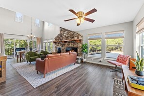 A spacious living room with a stone fireplace, a ceiling fan, and large windows offering a view of the outdoors. - 3 Kings! Hot Tub/Pool-Fire Pit-GameRm GYM+ PickleB (Canyon Lake)