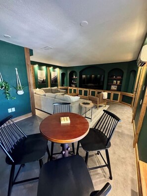 Une pièce avec une table ronde en bois et quatre chaises, un canapé et un téléviseur à l’arrière-plan.
