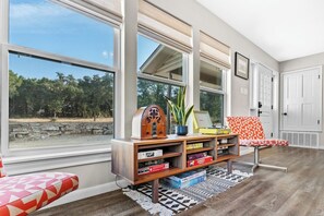 A room with large windows, a wooden entertainment unit, a red and white patterned bench, and a view of a stone wall and trees. - 3 Kings! Hot Tub/Pool-Fire Pit-GameRm GYM+ PickleB (Canyon Lake)