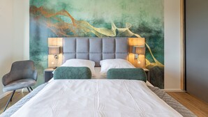 Quarto de hotel moderno com uma cama grande, duas mesas de cabeceira com luminárias, uma poltrona estofada verde e um grande mural abstrato na parede.