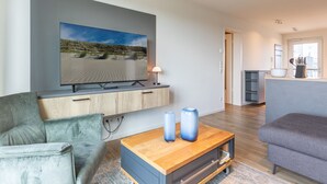 Uma sala de estar moderna com uma televisão de tela plana na parede, um sofá cinza, uma mesa de centro de madeira com dois vasos e um armário de madeira embaixo.