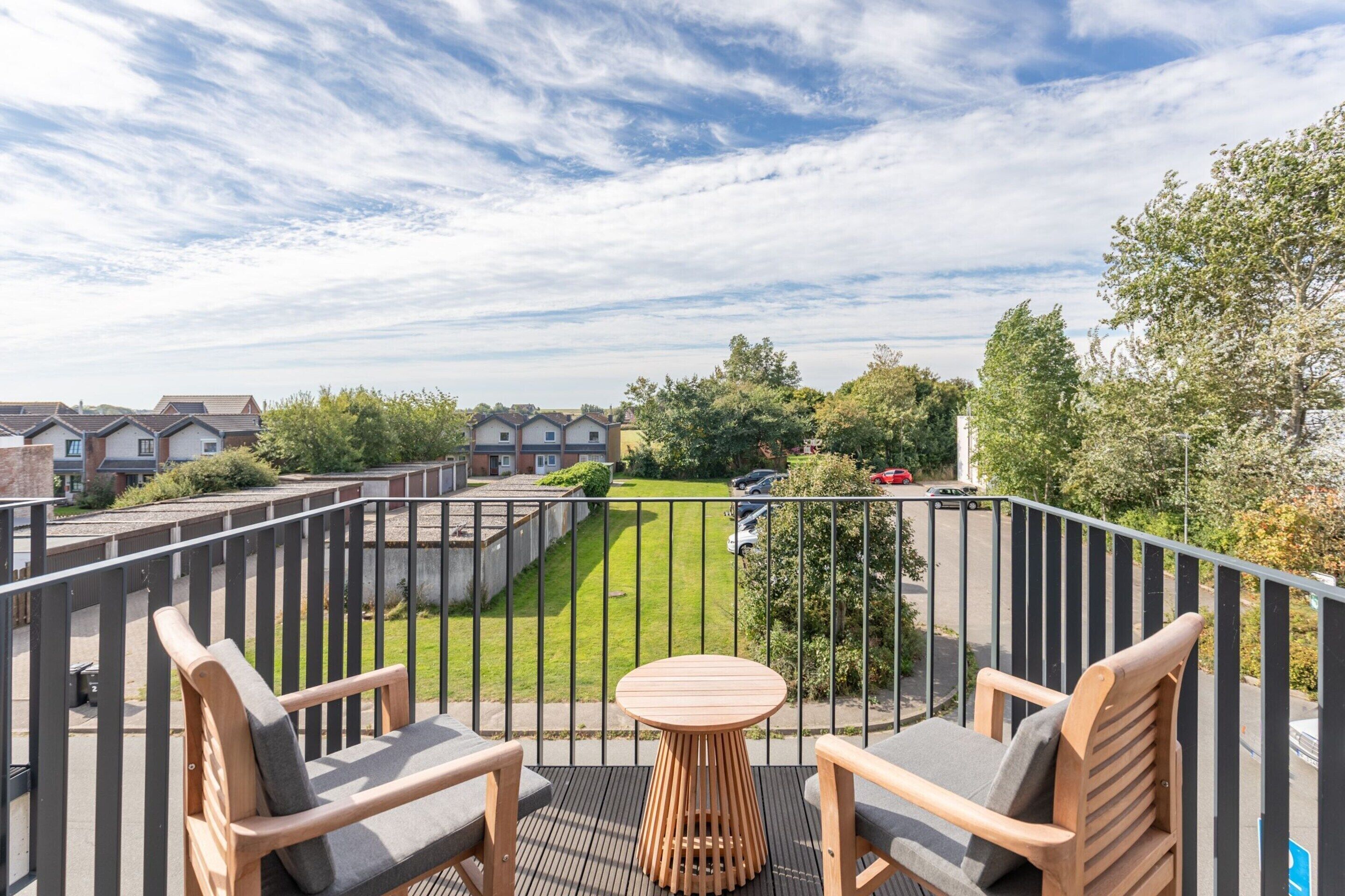 A balcony with a wooden table and two chairs, overlooking a well-maintained lawn and a parking area with cars.