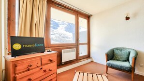 Une pièce avec une commode en bois, un téléviseur affichant l’émission « Maeva Home », une fenêtre donnant sur des montagnes enneigées et un fauteuil vert à pois.