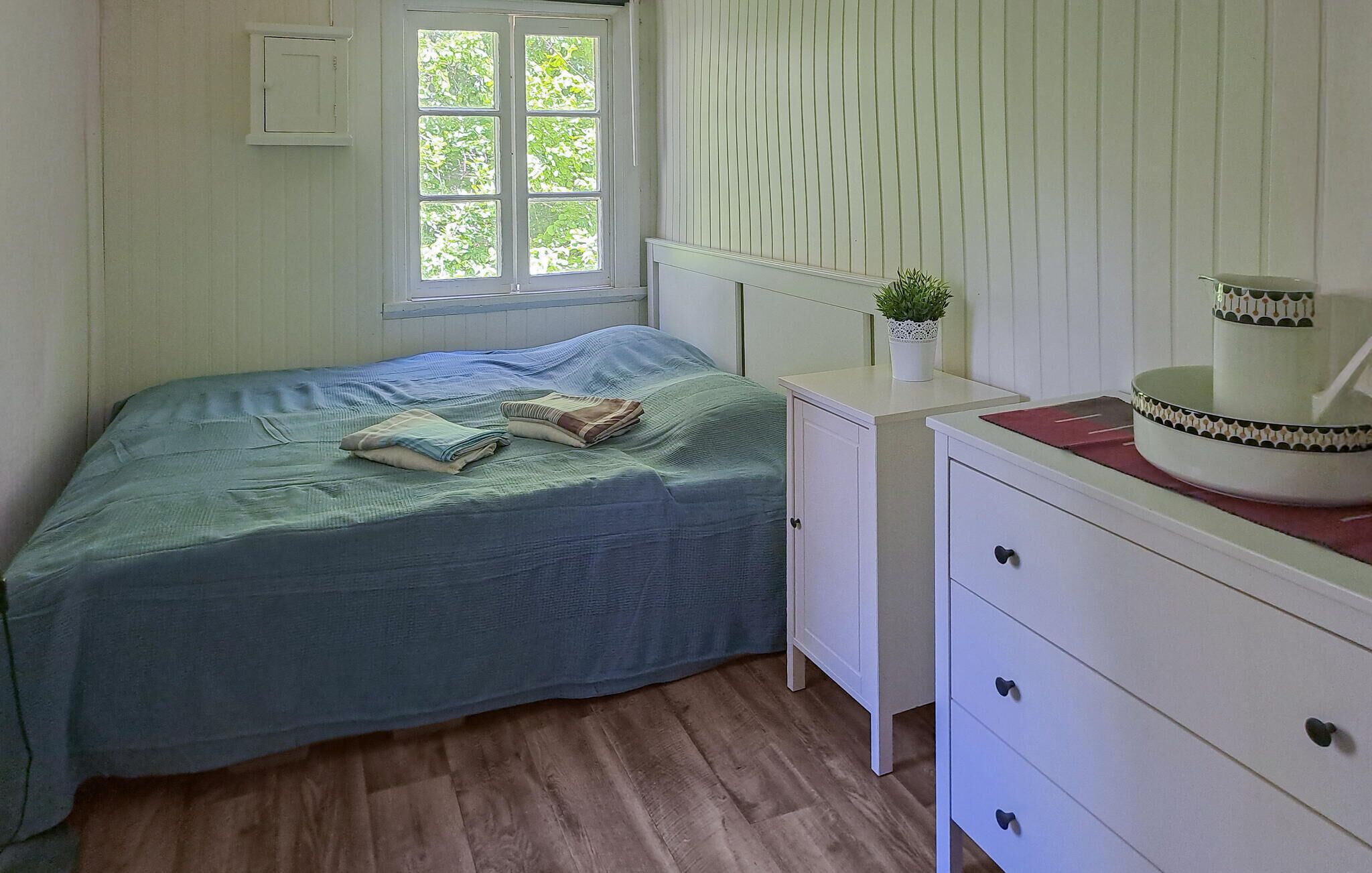 A bedroom with a bed, a bedside table with a plant, a dresser, and a window with blinds.