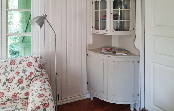 A room with a floral-patterned sofa, a floor lamp, a white cabinet with glass doors, and a window with a green shutter. - Amazing home in Halltorp with WiFi (Halltorp)
