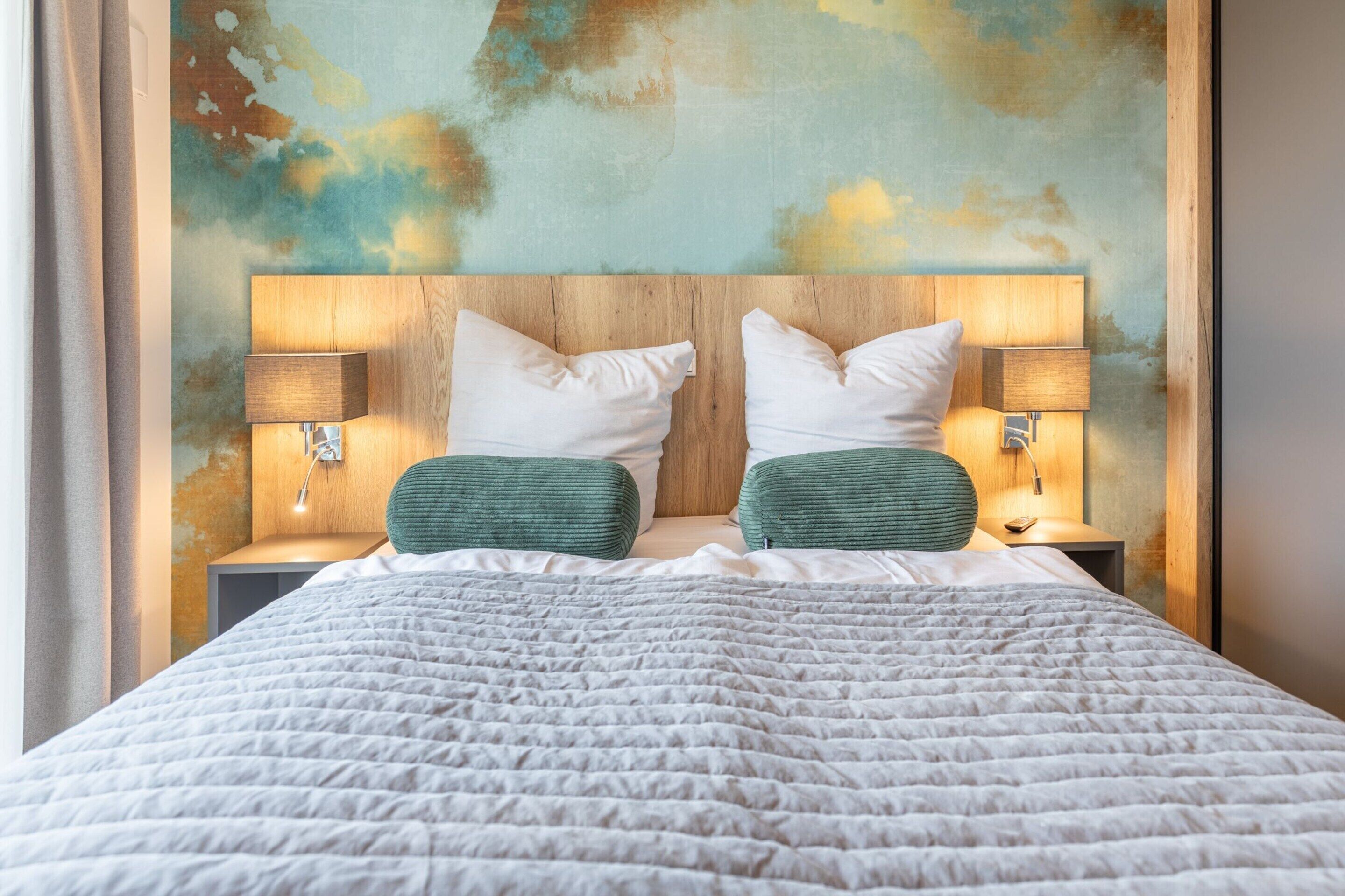 A neatly made bed with a quilted white duvet, two pillows, and two green accent pillows. There are two bedside lamps on wooden nightstands. The headboard is wooden with a textured finish.
