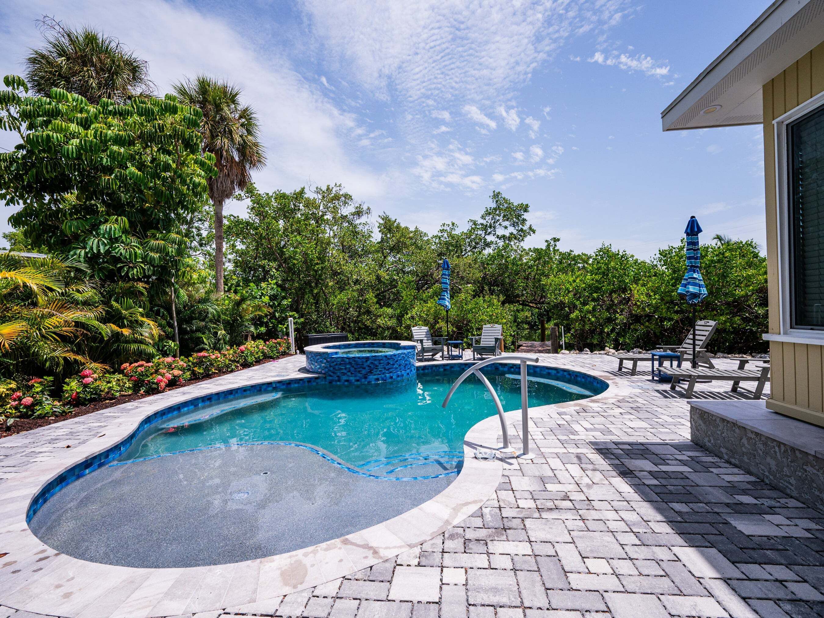 A swimming pool with a tiled deck, surrounded by lush greenery and a clear sky.