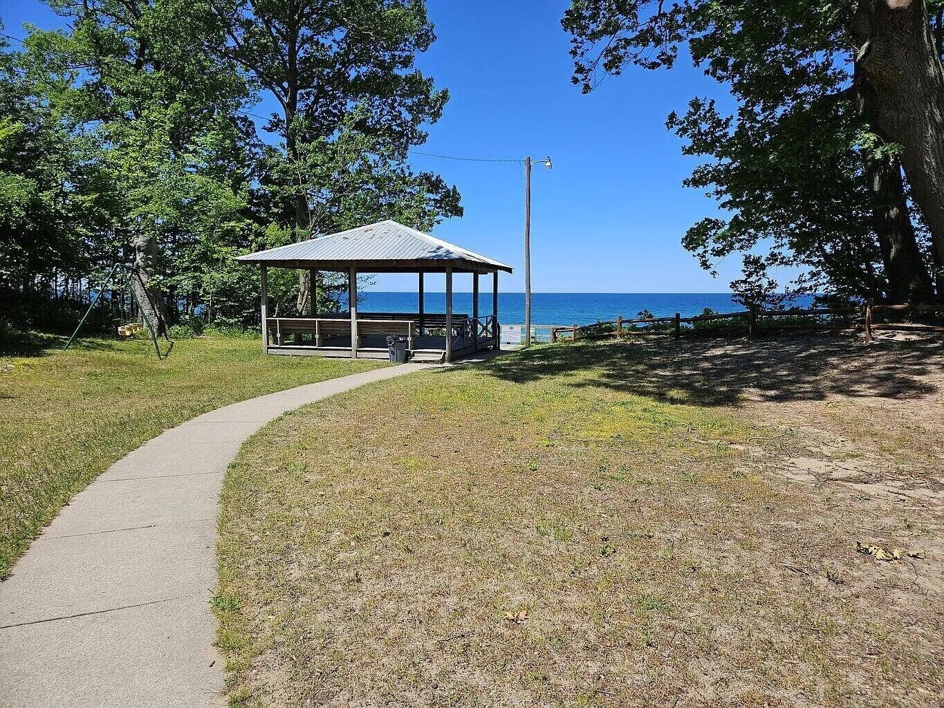 Un kiosque doté d’un toit en métal et de bancs en bois, entouré de pelouse et d’arbres, et offrant une vue sur une étendue d’eau.