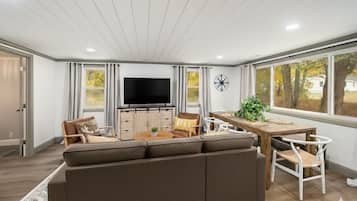 Una sala de estar moderna con un sofá modular, televisor, mesa de comedor con sillas y ventanas grandes con cortinas.