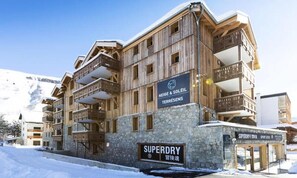 Un edificio de madera de varios pisos con balcones, identificado con las etiquetas "NEIGE & SOLEIL TERRÈSES" y "SUPERDRY".