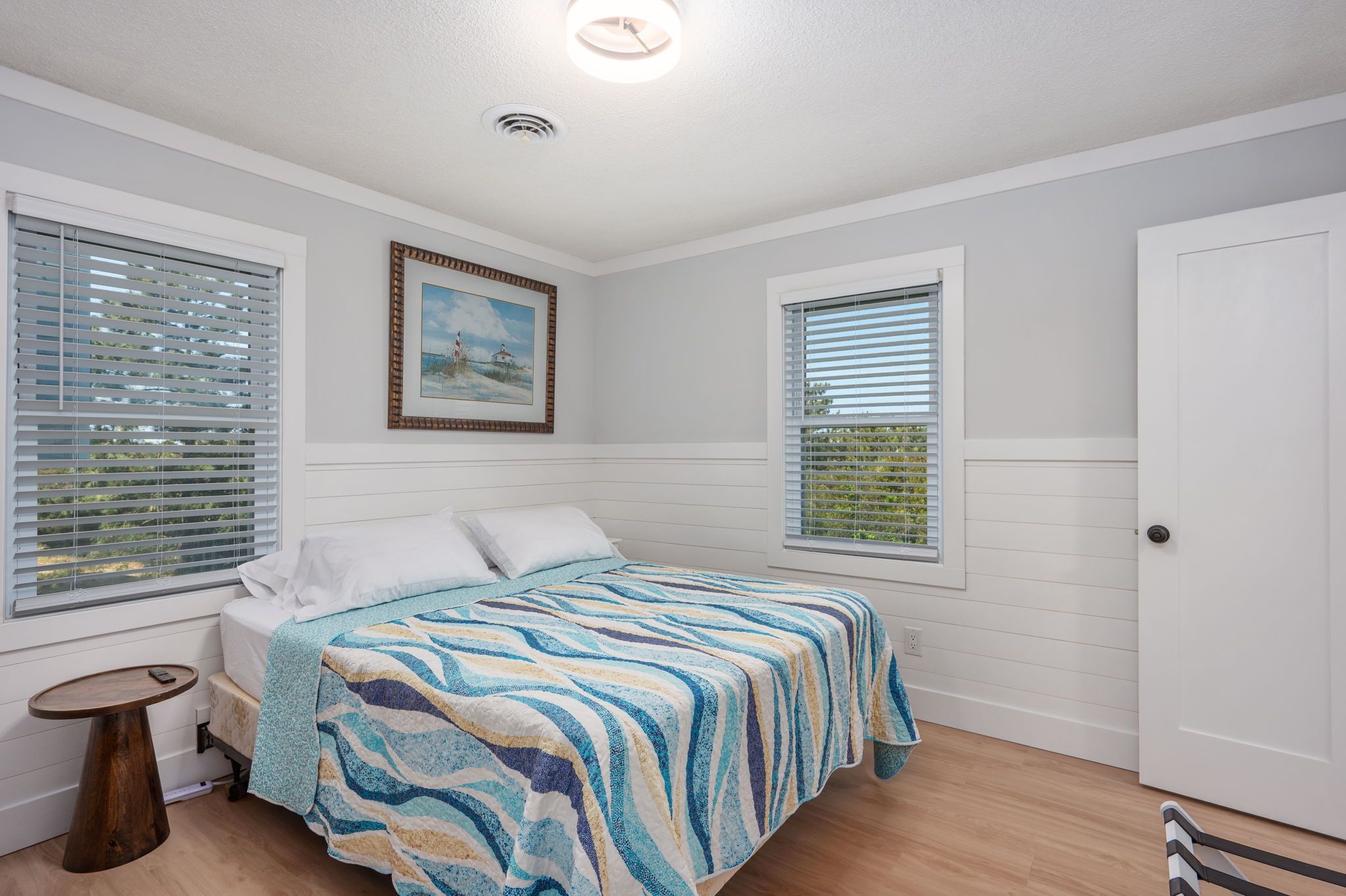 A bedroom with a bed, two windows with blinds, a painting on the wall, and a door.