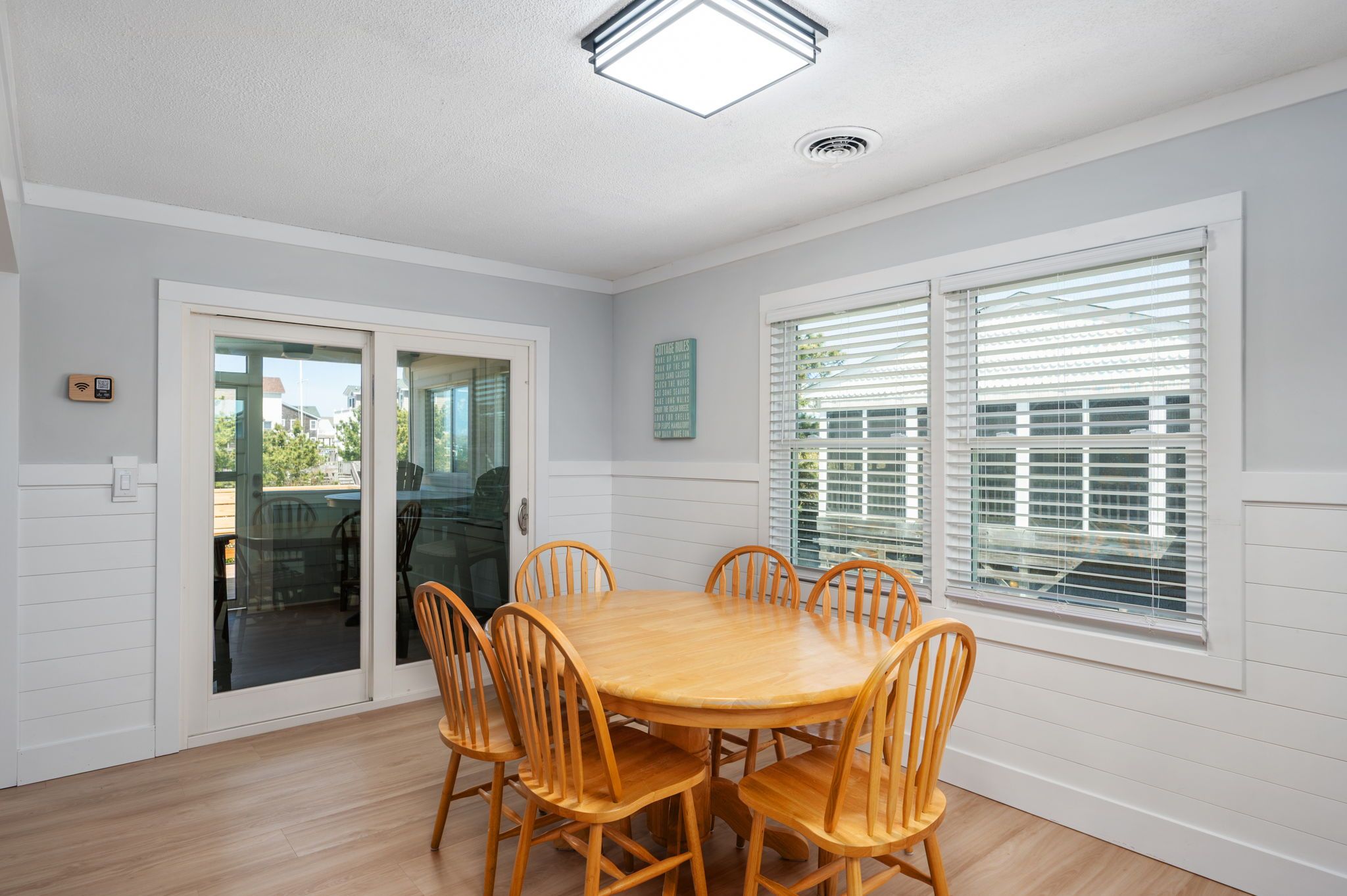 A dining area with a round wooden table and six chairs, a sliding glass door, and a window with blinds.