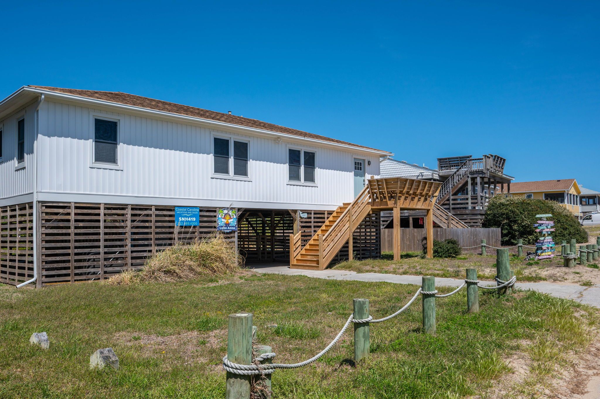 A coastal building with a wooden staircase leading to a covered area, surrounded by a grassy area with a fence.