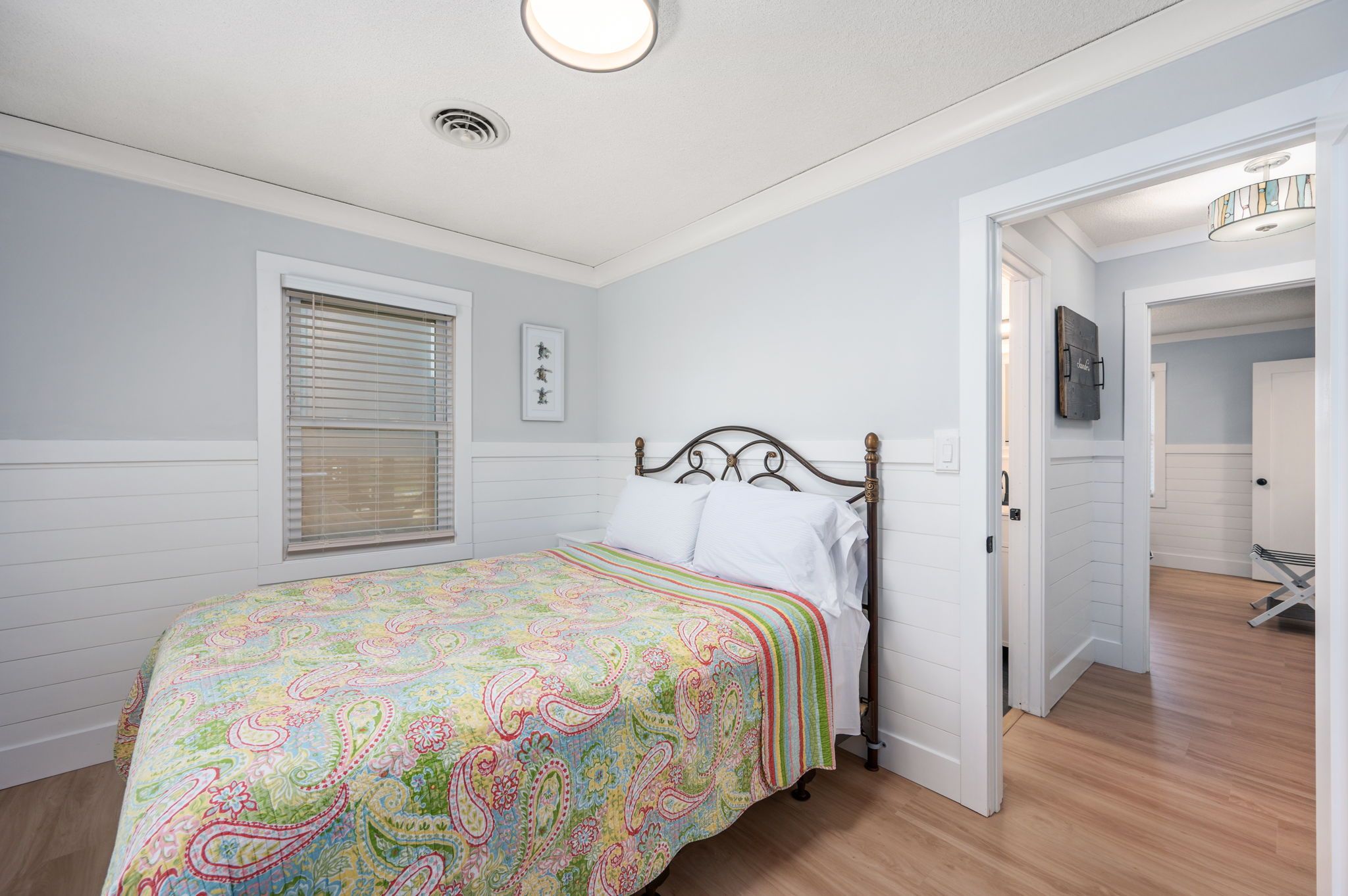 Une chambre à coucher avec un lit, une fenêtre avec des stores, une porte donnant sur une autre pièce et un luminaire au plafond.