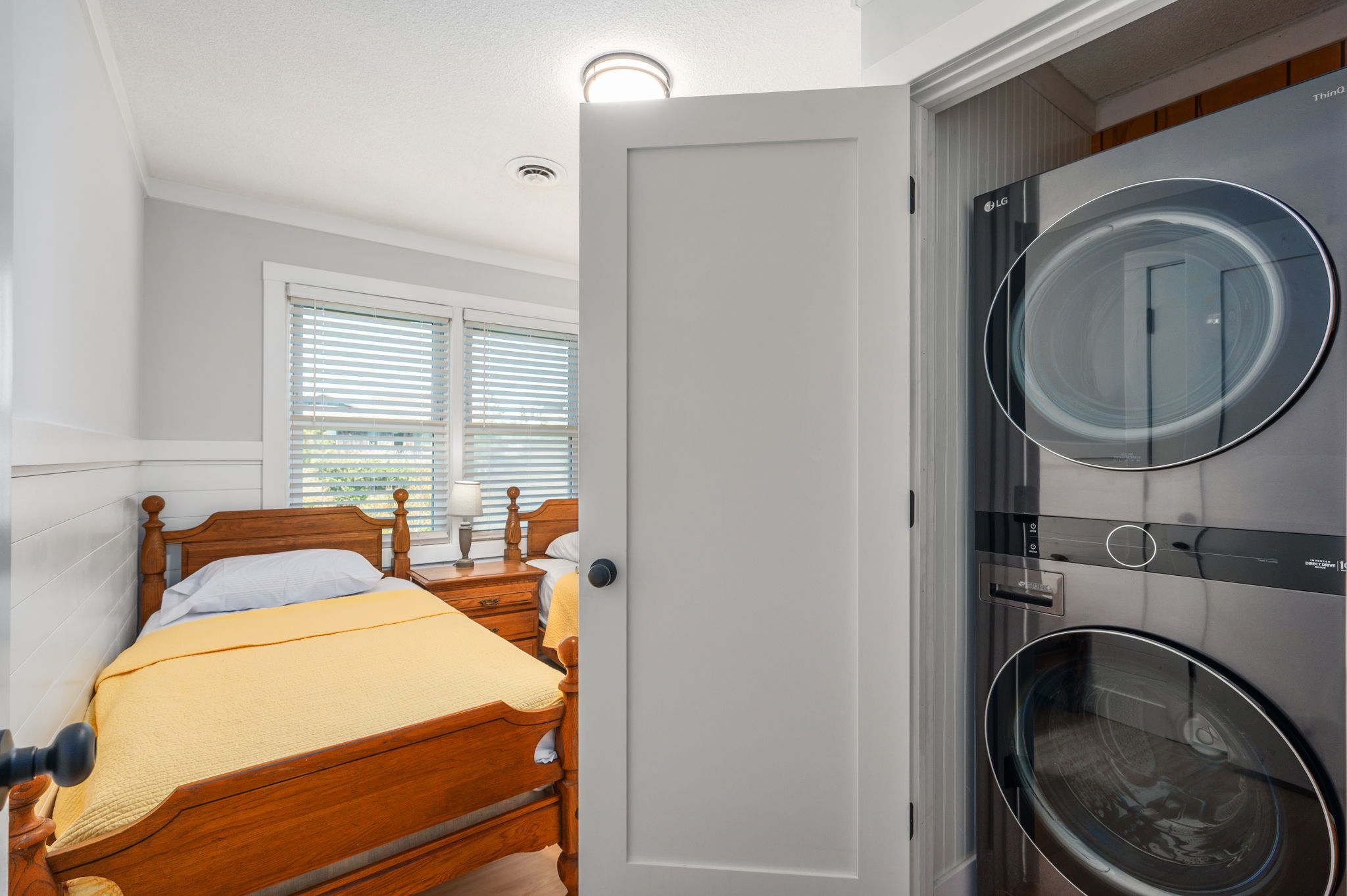 A bedroom with a wooden bed, a nightstand, a lamp, and a door leading to a laundry area with two washing machines.