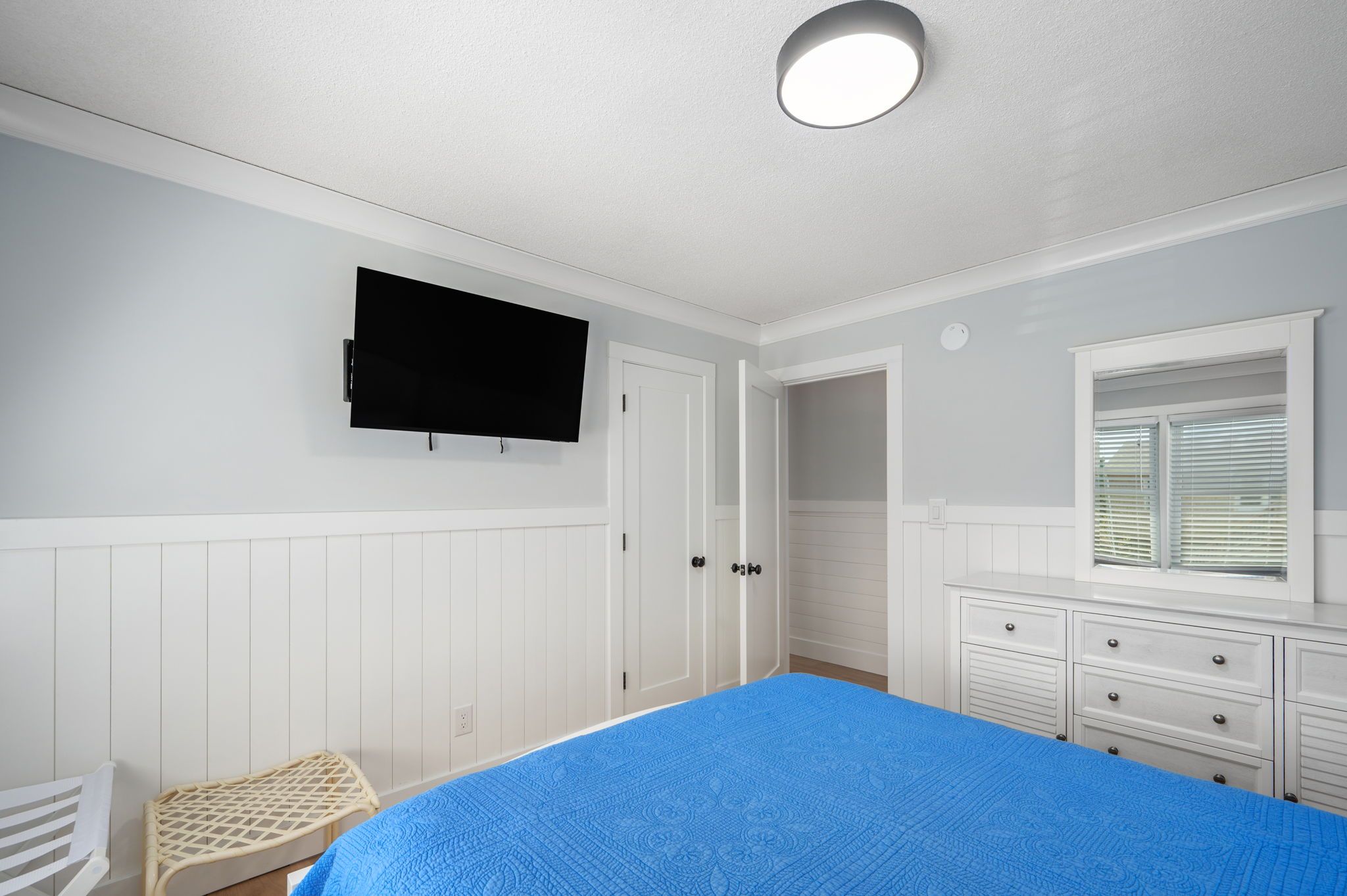 Une chambre à coucher comprenant un lit, un téléviseur fixé au mur, une commode et une fenêtre avec des stores.