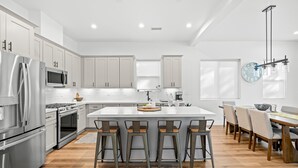 A modern kitchen with stainless steel appliances, a central island with bar stools, and a dining area with a long table and chairs.