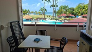 A balcony with a table and chairs, a view of a pool and beach, and a building with red roofs. - Building Martinique (Sainte-Luce)