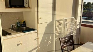 A small kitchen with a microwave, sink, and cabinets. There is a chair and a table with a view of palm trees outside. - Building Martinique (Sainte-Luce)