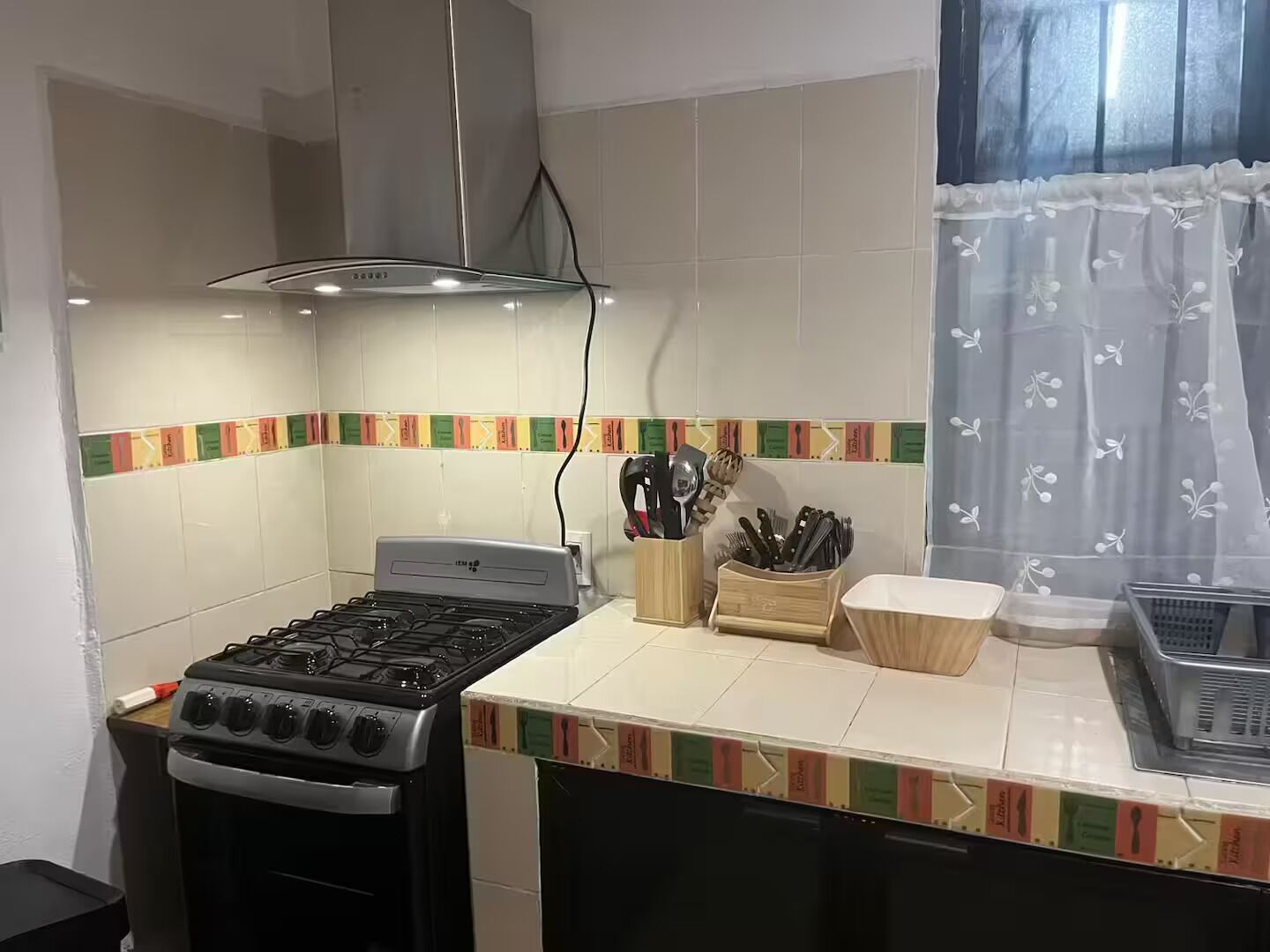 Une cuisine équipée d’une cuisinière à gaz, d’une hotte aspirante et d’une fenêtre ornée d’un rideau léger.